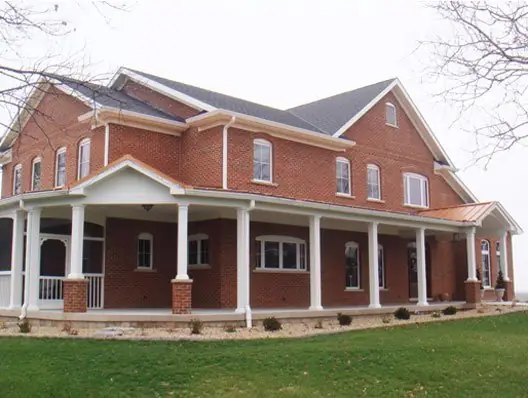 Two storey brick home built by C.B.E. Construction with a white wraparound porch, columns and a metal porch roof